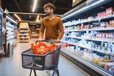 Genç adam süpermarkette bir alışveriş arabasını itiyor, temiz ve parlak koridorlardaki raflardan taze sebzeler seçiyor.