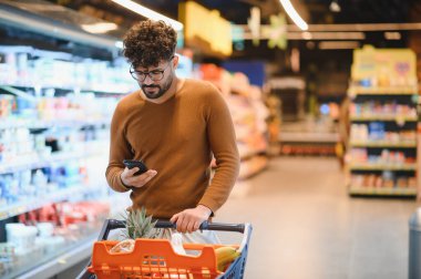 Genç adam alışveriş listesini kontrol ederken marketin içinde alışveriş arabasıyla geziyor.