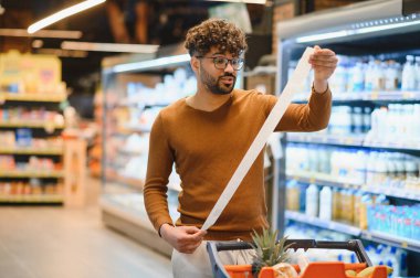 Markette alışveriş yaparken çok uzun bir makbuzu inceleyen müşteri, ürünlerin yüksek maliyetine şaşırır.