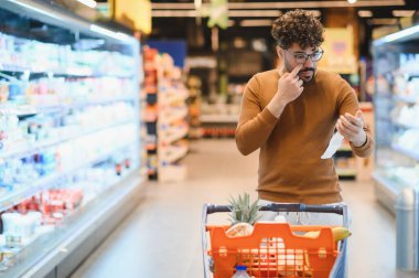 Arap müşteri alışveriş arabasını itiyor, uzun bir makbuzu kontrol ediyor ve markette alışveriş yaparken burnuna dokunuyor.
