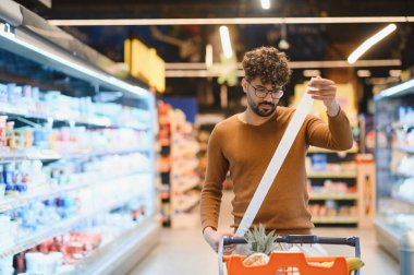 Genç adam, süpermarkette alışveriş yaparken uzun bir market makbuzunu dikkatle inceleyerek, enflasyonun tüketici harcamaları ve bütçeleme üzerindeki etkisini vurguluyor
