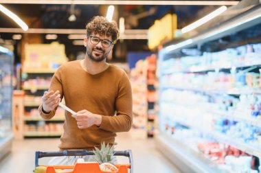 Genç adam alışveriş listesini kontrol ederken elinde alışveriş arabasıyla süpermarket koridorlarında yürüyor.