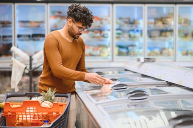Genç Arap adam markette alışveriş yapıyor, dondurulmuş yiyecekleri soğuk bir koridordan seçiyor.