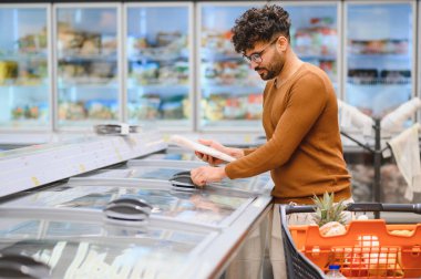 Arap adam market alışverişi yapıyor, marketteki dondurulmuş yiyecekleri seçiyor, elinde dondurulmuş bir ürün paketi tutuyor.