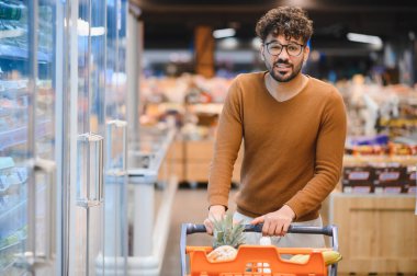 Genç Arap adam süpermarkette alışveriş arabasını itiyor market alışverişi yapıyor, dondurulmuş bölümde yürüyor.