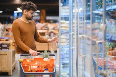 Müşteri bir süpermarkette buzdolabının kapısını açıyor, taze yiyecek ararken parlak koridorda bir alışveriş arabasını itiyor.