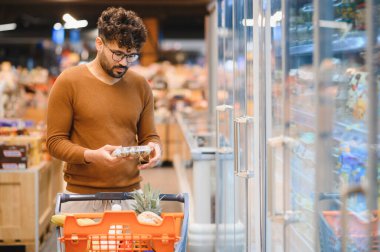 Genç Arap bir adam market buzdolabından dondurulmuş yiyecek seçerken alışveriş arabasını itiyor, günlük alışverişle meşgul.