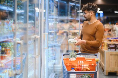 Genç adam market alışverişi yapıyor, alışveriş arabası itiyor ve dondurulmuş ürünleri süpermarketteki dondurulmuş koridordan seçiyor.