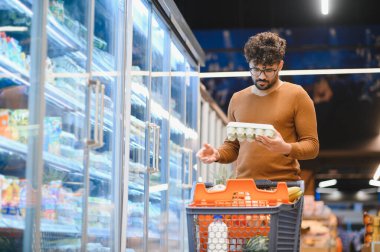 Müşteri, marketteki soğutuculu koridordan taze yumurta seçerken içi yiyecek dolu bir alışveriş arabasını itiyor.