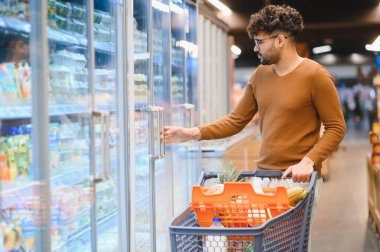 Genç Arap adam süpermarkette buzdolabı açarken market alışverişi yapıyor, alışveriş arabası itiyor.