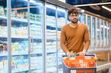 Genç bir adam, her hafta yediği şeyler için raflardan taze sebze seçen parlak, düzenli bir süpermarkette alışveriş arabasını itiyor.