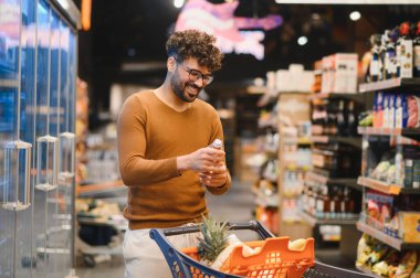 Genç Arap adam gülümsüyor ve süpermarkette bir şişe su seçiyor, alışveriş arabasını poşetlerle itiyor.