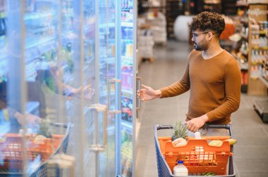 Genç adam alışveriş arabasını itiyor ve buzdolabının kapısını açıyor. Süpermarketten alışveriş yapmayı seçiyor.