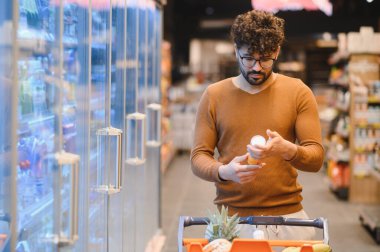 Parlak, modern bir süpermarkette market alışverişi yaparken ürün malzemelerini inceleyen genç adam sağlıklı bir yaşam tarzı için dikkatli seçimler yapıyor.