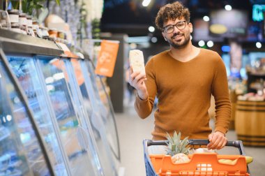 Genç Arap adam alışveriş arabasını itiyor ve markette soğutulmuş rafların yanında peynir gösteriyor.