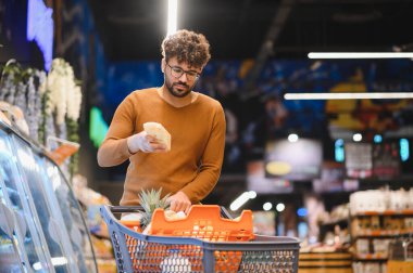 Müşteri market alışverişi yapıyor, elinde bir paket pide ekmeği tutuyor, malzemeleri kontrol ediyor, markette alışveriş arabasına yemek koyuyor.