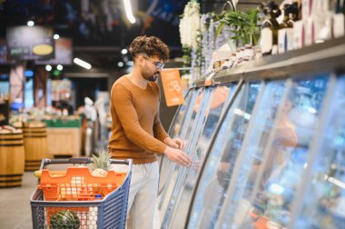 Müşteri, parlak ışıklı bir süpermarkette soğutuculu vitrinlerden sebze seçiyor, alışveriş arabasını itiyor.