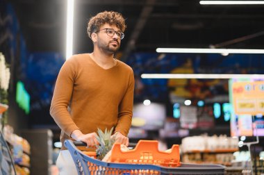 Genç yetişkin erkek süpermarkette alışveriş arabasını itiyor, market alışverişi yapıyor, parlak, modern markette sağlıklı yiyecekleri seçiyor.
