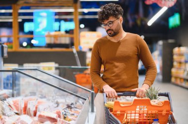 Arap adam alışveriş arabasını itiyor ve dondurulmuş ürünleri marketin dondurulmuş bölümünde seçiyor.