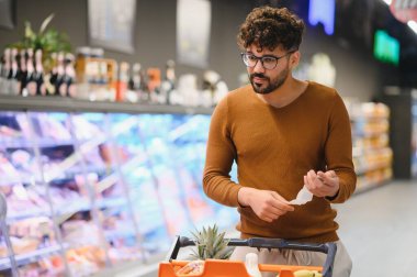 Genç Arap adam alışveriş listesini kontrol ederken bir süpermarkette alışveriş arabasıyla alışveriş yapıyor.