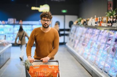 Genç Arap bir adam süpermarkette alışveriş arabasını itiyor, taze sebzeler seçiyor ve kalabalık bir mağazanın koridorlarında geziniyor.