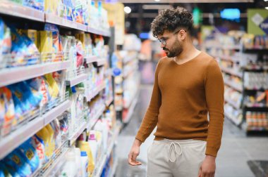 Genç Arap adam deterjan alışverişi yapıyor. Parlak bir süpermarket koridorunda etiketleri dikkatle okuyor.