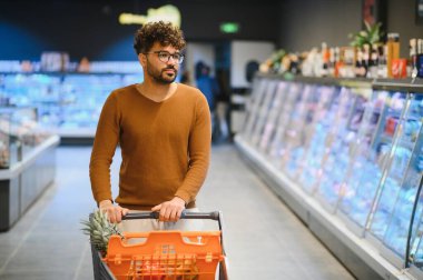 Genç Arap bir adam alışveriş arabasını iterken aynı zamanda çeşitli ürünlerle dolu, ferah bir süpermarket koridorunda taze yiyecekler seçiyor.