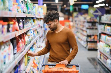 Genç Arap adam, parlak ışıklı bir süpermarkette alışveriş yaparken alışveriş yapıyor, çeşitli ürünlerle dolu koridorlarda geziniyor.