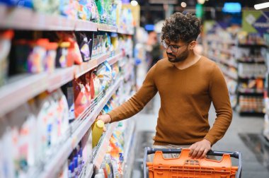 Genç Arap adam raftan ürün topluyor, süpermarkette alışveriş yapıyor ve alışveriş arabasıyla alışveriş yapıyor.