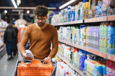 Genç Arap bir adam alışveriş arabasını iterken aynı zamanda çeşitli mallarla dolu bir süpermarket koridorunda temizlik ürünleri seçiyor.