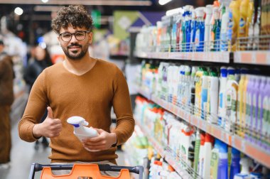 Genç adam elinde temizlik malzemesi tutarken başparmağını kaldırıyor, çeşitli malzemelerle dolu canlı bir süpermarkette alışveriş yapıyor.