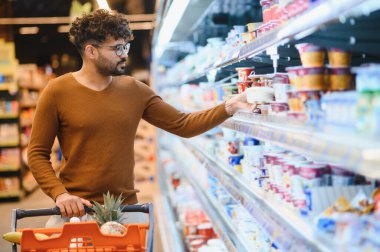 Genç bir adam alışveriş arabasını iterken buzdolabı bölümünden yoğun bir süpermarket koridorunda yoğurt seçiyor.