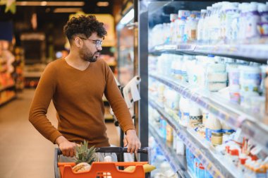 Müşteri alışveriş arabasını itiyor ve modern süpermarketteki soğutulmuş raflardan taze süt ürünleri seçiyor.