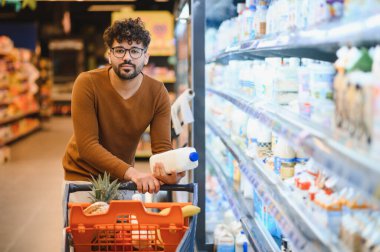 Müşteri bakkal alışverişi yapıyor, elinde bir şişe süt tutuyor süpermarketin buzdolabı bölümünde alışveriş arabasının yanında duruyor.