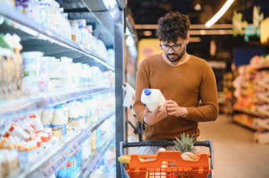 Odaklanmış müşteri süt etiketi okuyor, süpermarket koridorunda alışveriş arabasıyla satın alma kararı veriyor.