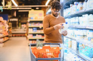 Genç Arap adam marketin buzdolabı koridorunda alışveriş yaparken süt şişesi etiketini dikkatlice okuyor, alışveriş arabasını taze ürünlerle itiyor.