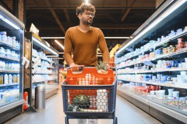 Genç Arap adam süpermarket koridorunda yürürken alışveriş arabasını itiyor.