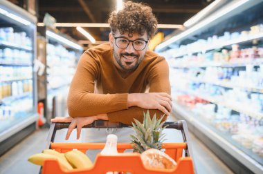 Genç Arap adam, canlı bir süpermarket koridorunda alışveriş arabasına yaslanırken gülümsüyor.