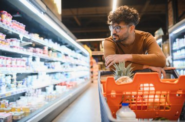 Genç Arap bir adam alışveriş arabasına yaslanmış taze ürünlerle dolu, parlak, düzenli bir süpermarket koridorunda yiyecek seçiyor.
