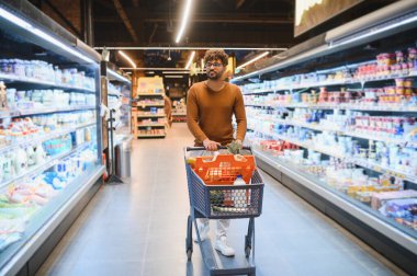 Genç bir adam market koridorunda alışveriş arabasıyla dolu bir alışveriş arabasını itiyor ve rutin bir alışveriş deneyiminin tadını çıkarıyor.