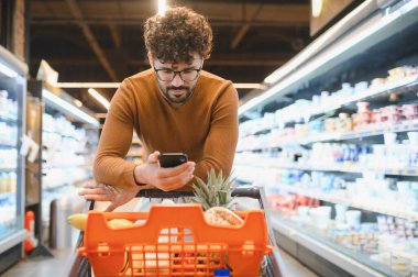 Genç adam alışveriş listesini akıllı telefondan kontrol ederken marketin kalabalık koridorunda taze yiyecek seçiyor.