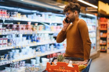 Müşteri, raflardan gıda ürünleri seçerken, marketlerde alışveriş arabalarını iterken, cep telefonuyla konuşuyor.