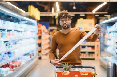 Alışverişçinin, markette alışveriş arabasını iterken çok uzun bir makbuzu incelemesi, marketin yüksek maliyetine şaşırır.