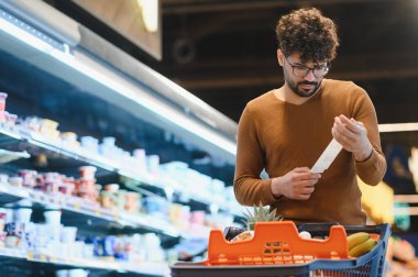 Genç adam süpermarkette alışveriş yaparken market fişini kontrol ediyor. Bütçesine uyduğundan emin olmak için.