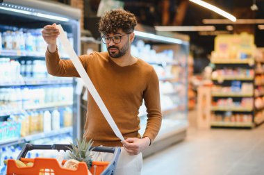 Müşteri, süpermarket koridorlarında gezinirken, harcamaları yönetirken ve gıda alımları için bütçe ayırırken uzun bir market fişini kontrol ediyor