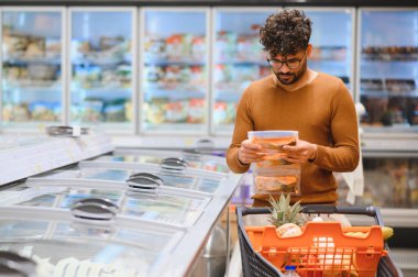 Genç adam marketin dondurulmuş gıda reyonunda alışveriş yaparken yiyecek etiketini dikkatlice okuyor.