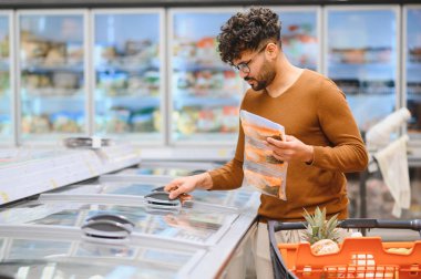 Market alışverişi yapan, süpermarket koridorunda buzdolabı açan, dondurulmuş gıda paketi tutan genç bir adam.