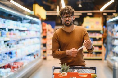 Genç adam süpermarkette alışveriş yaparken market fişini kontrol ediyor. Bütçeyi aşmadığından emin oluyor.