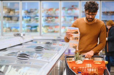 Adam market alışverişi yapıyor, dondurulmuş ürünlerin malzemelerini süpermarket buzdolabı koridorunda kontrol ediyor, alışveriş arabasını itiyor.