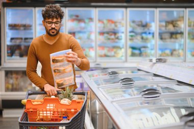 Genç adam market alışverişi yapıyor, markette dondurulmuş balık filetosu seçiyor.